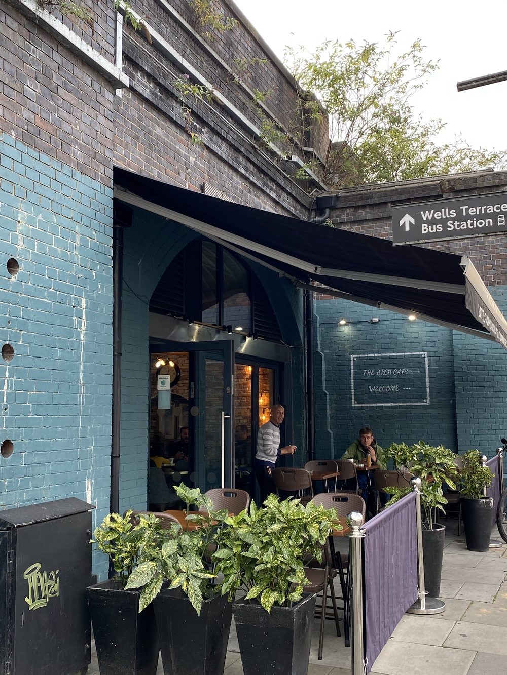 The teal-painted brick arch shopfront of Arch Cafe with awning, planters and pavement seating on Wells Terrace
